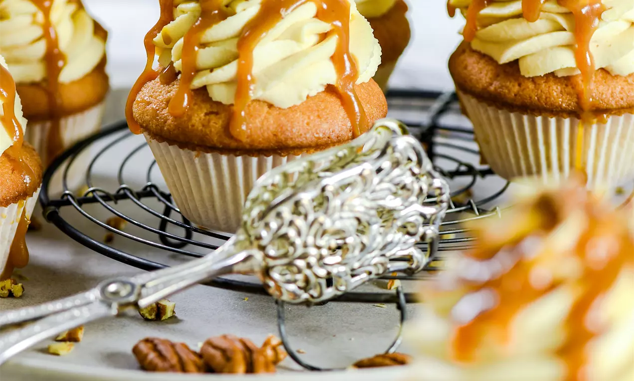 Picture - Karamell och pecan cupcakes med gyllene glasyr, spritsad grädde och hackade nötter på ett galler med ett dekorativt bakverktyg